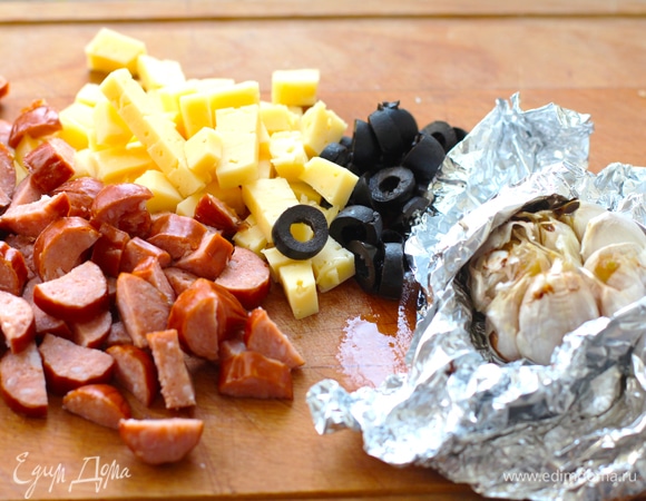 Чеснок запекать в фольге в духовке около 20 минут при 170°C (верхушку немного срезать и влить оливковое масло). Остудить, раздавить вилкой. Все добавки нарезать. Сыр нужен твердый, но плавкий, пармезан не подойдет. Оливки нарезать кольцами, копчености —кубиками.