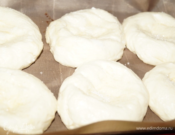 Разделите тесто на шарики, если оно будет немного прилипать, смажьте руки растительным маслом. Шарики выложите на противни, застеленные пекарской бумагой или ковриком для выпечки. Сформируйте основу для ватрушки из шарика, надавив ложкой на середину, превратив шарик в лепешку с углублением по середине. В углубление насыпьте сахарный песок, разровняйте его ложкой.