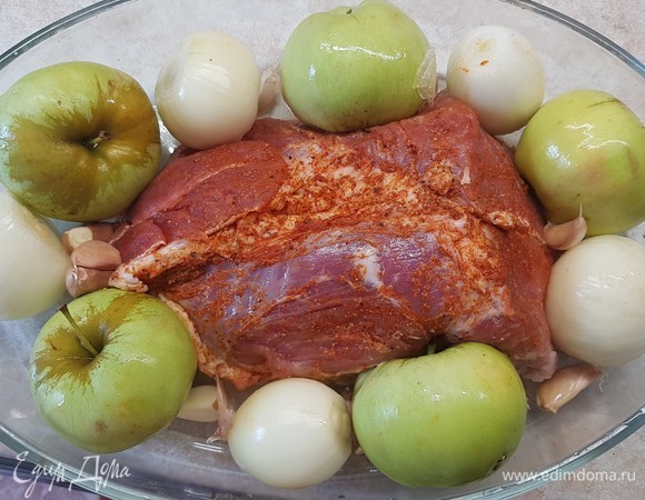 Мясо уложить в форму для запекания. Вокруг куска выложить целые яблоки и луковицы. Чеснок разобрать на зубчики, но не чистить их. Выложить тоже вокруг свинины.