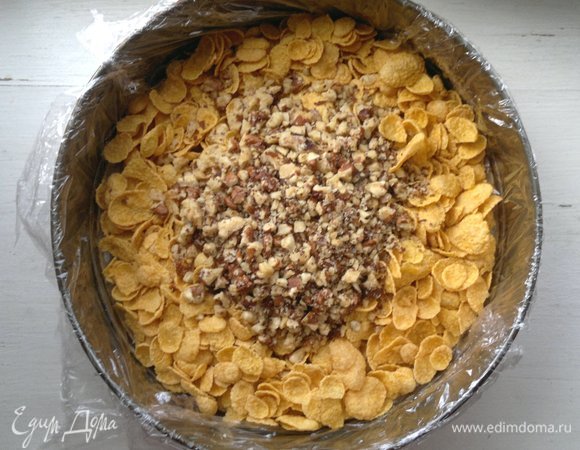 Разъемную форму для выпечки застелить пищевой пленкой. Выложить грецкие орехи и кукурузные хлопья. Как потом выяснилось, форму лучше застилать фольгой, так как через пленку немного вытекала карамель, пока не застыла. У меня форма диаметром 26 см. Конечно, в форме меньшего диаметра торт получился бы выше.