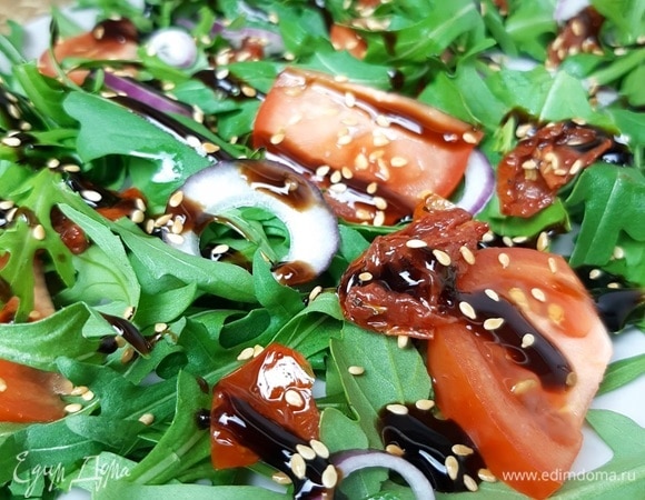 На сервировочную тарелку выкладываем листья руколы. Если они слишком крупные, можно их порвать руками. Сливаем воду с красного лука и выкладываем его на зелень. Далее — помидоры и вяленые томаты. Всю эту красоту поливаем бальзамическим соусом и посыпаем семенами ароматного кунжута. Приятного аппетита!