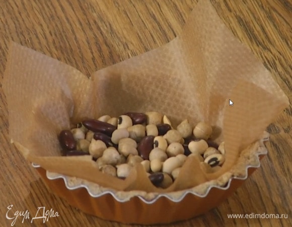 Охлажденное тесто распределить по дну и стенкам формочек для тарталеток, вложить в каждую пекарскую бумагу, сверху насыпать фасоль или бобы.