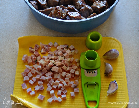 Тем временем бекон нарежьте мелкими кубиками. Чеснок почистите и измельчите с помощью чеснокодавилки. Поместите бекон в сотейник, в котором обжаривалось мясо, и слегка поджарьте. Добавьте туда же измельченный чеснок и готовьте 2 мин, перемешивая.