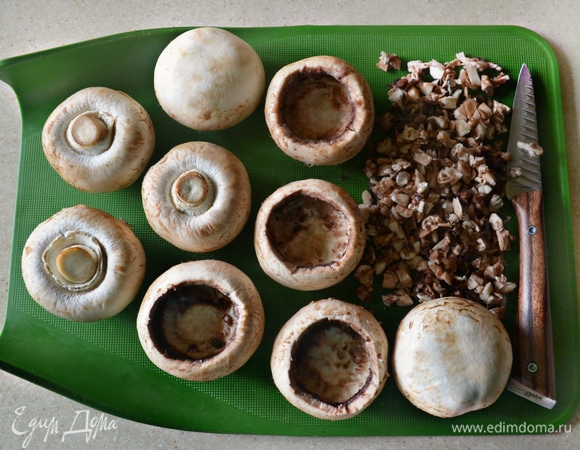 Шампиньоны (нам понадобятся достаточно крупные грибы) промойте, просушите и почистите. Острым ножом аккуратно вырежьте ножки и верхнюю часть шляпок. Вырезанные части грибов мелко порубите. Поместите очищенные шапки шампиньонов в форму для запекания, смазанную растительным маслом. Сверху немного сбрызните грибы маслом, накройте фольгой и запекайте в разогретой до 180°C духовке 20 мин.