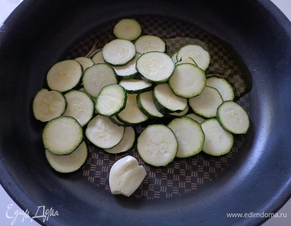 Цукини помыть, нарезать. В сковороду налить оливковое масло, добавить зубчик чеснока и обжарить цукини (3–4 минуты).