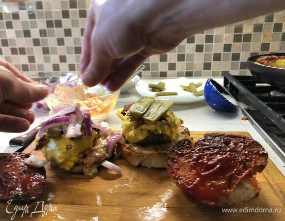 На нижнюю булочку выкладываем соус с салатом, далее — котлета в сыре с беконом, соленый огурчик, лук. Верхнюю булочку смазываем кетчупом и ей закрываем.