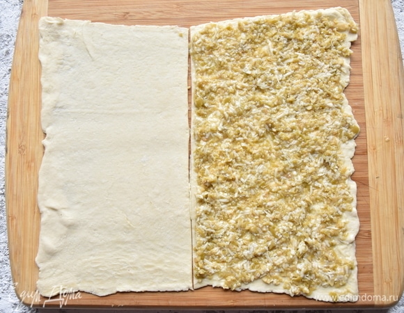 Слоеное тесто раскатать в прямоугольник и разрезать его на две равные части. На одну из них выложить подготовленную начинку и распределить по всей поверхности.