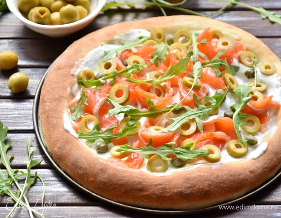 Выпекать тесто 12 минут в разогретой до 250°C духовке. Смазать центр горячей лепешки сметаной. Выложить ломтики лосося, оливки, каперсы и руколу.