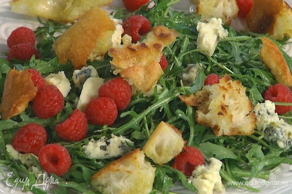 Выложить заправленную руколу на большую тарелку, сверху разложить кусочки сыра, добавить ягоды малины и крутоны.