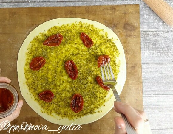 Припыляем стол мукой. Из шарика теста руками аккуратно формируем небольшую круглую лепешку. Затем раскатываем ее в круг диаметром примерно 23 см. Тогда фокачча получится тоненькой и хрустящей. Перекладываем тесто на коврик или пергамент, стряхнув лишнюю муку. Смазываем края теста оливковым маслом. Наносим на фокаччу соус песто и выкладываем вяленые томаты.