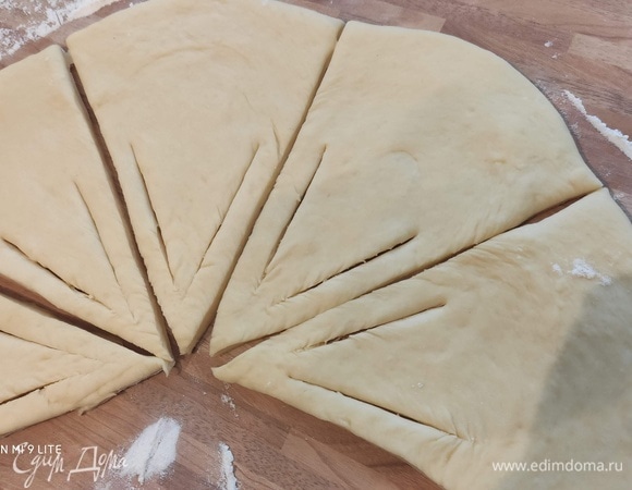 Расстоявшееся тесто необходимо обмять, разделить на две части, раскатать в круг диаметром ~ 40 см. Каждый пласт теста разрезать острым ножом на восемь частей. Надрезать каждый треугольник на кончике (по одному надрезу слева и справа) вдоль длинной стороны примерно на 5 см и 1 см от верхушки треугольника. Совет: пожалуйста, убедитесь, что кончики теста не прорезаны.