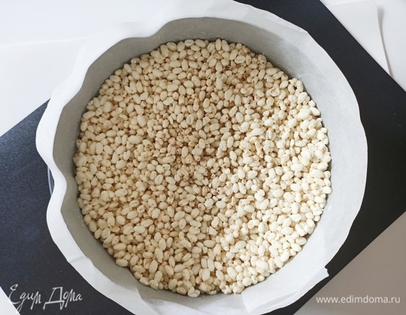 Белый шоколад (150 г) необходимо растопить на водяной бане и смешать с воздушным рисом. Форму для выпечки (22 см) застелить пергаментной бумагой. На дно формы выкладываем рисовые шарики с шоколадом и отправляем форму в холодильник на 30 минут.