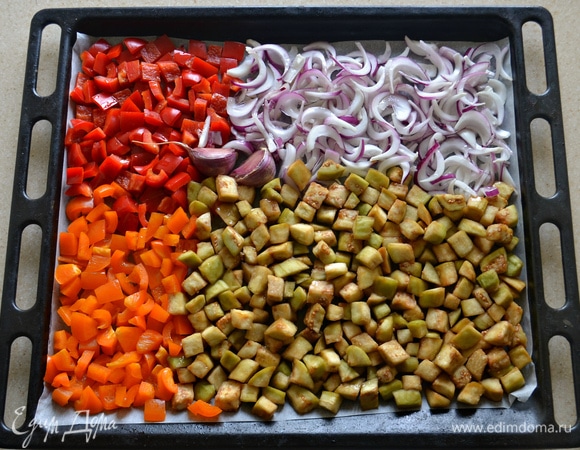 Перец очистить от семян и нарезать кубиками. Лук очистить и нарезать перьями. Уложить овощи вместе с неочищенными зубчиками чеснока на противень, застеленный пекарской бумагой. Сбрызнуть оливковым маслом и запекать при 180°C около 40 минут, пока овощи не станут мягкими. Во время запекания овощи пару раз нужно аккуратно перемешать.
