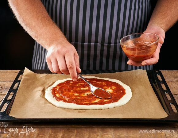 Раскатайте тесто в круглую форму, сформировав небольшие бортики, выложите на противень, застеленный пергаментом. Смажьте основу томатным соусом.