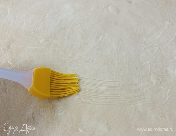 Как только тесто поднимется, припыляем поверхность стола мукой и раскатываем его скалкой в прямоугольник. Чем тоньше раскатаем, тем больше будет слоев. Сильно размягченным сливочным маслом смазываем раскатанное тесто, кроме одноного из краев по длинной стороне шириной приблизительно 2 см.