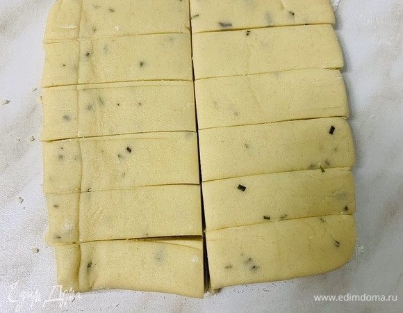 Руками замешиваем гладкое тесто. Делим пополам и раскатываем одну половину на слегка припыленной мукой рабочей поверхности в квадрат размером 12 х 12 см и толщиной около 1 см. Нарезаем на шесть полосок шириной 2 см, затем разрезаем каждую полоску, чтобы получилось 12 печенек.