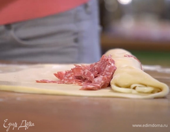 На присыпанной оставшейся мукой рабочей поверхности раскатать из подошедшего теста две тонкие продолговатые лепешки, на край длинной стороны каждой лепешки выложить сулугуни, завернуть немного тесто с сыром к центру, затем выложить салями и свернуть все в рулет.