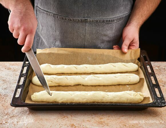 Разогрейте духовку до 220°С. Противень застелите пергаментной бумагой. Тесто разделите на три части и сформируйте багеты. С помощью ножа сделайте неглубокие надрезы на заготовках, положите их на противень и оставьте подойти на 10 минут.