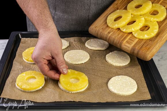 Раскатайте тесто толщиной 4 мм. При помощи кружки или кулинарного кольца диаметром равным диаметру ананаса сделайте круглые заготовки. Застелите противень бумагой для выпекания, выложите кружки из теста, на них поместите колечки ананасов.