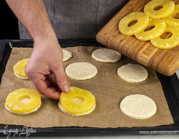 Раскатайте тесто толщиной 4 мм. При помощи кружки или кулинарного кольца диаметром равным диаметру ананаса сделайте круглые заготовки. Застелите противень бумагой для выпекания, выложите кружки из теста, на них поместите колечки ананасов.