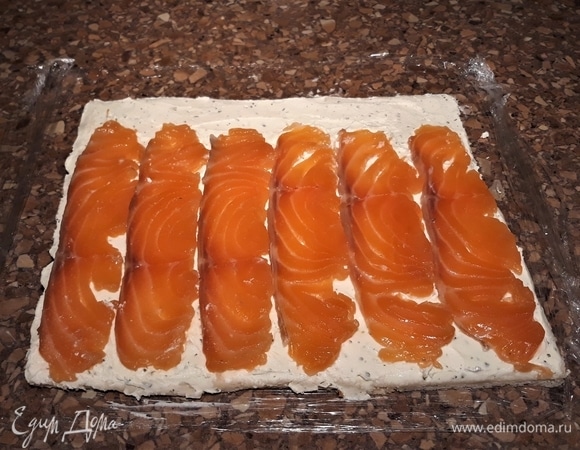 На пищевую пленку уложите хлеб слегка внахлест по три кусочка в два ряда. Скалкой примните, чтобы не было швов. Смажьте хлебный пласт творожно-сливочным сыром. Сверху выложите полоски рыбы.