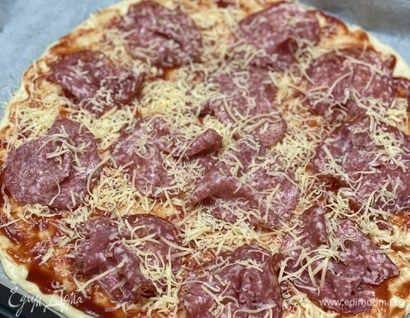 Духовку разогреть до 200°C. Противень застелить пергаментом, выложить на него половину теста и руками растянуть до диаметра 30 см, придавая форму круга. Смазать основу соусом, посыпать тертым сыром, разложить колбасу (я использую только в нарезке, так кусочки максимально тонкие), далее — помидоры, сверху — еще немного сыра.