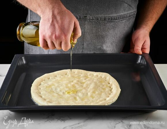 Подошедшее тесто разделите на две части, одну из которых раскатайте в пласт толщиной 2–3 мм. Оставшуюся часть теста можно использовать для второй порции.<br />
Переложите раскатанное тесто на противень, сбрызните оливковым маслом.