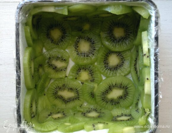Квадратную форму (у меня размер — 12 х 12 х 6) застелить пищевой пленкой, выложить дно и бока кружками киви.