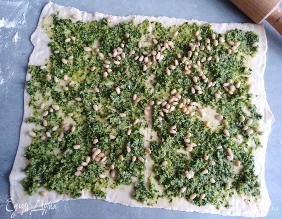 Тесто заранее разморозить. Раскатать в пласт. Щедро смазать песто, посыпать орешками.