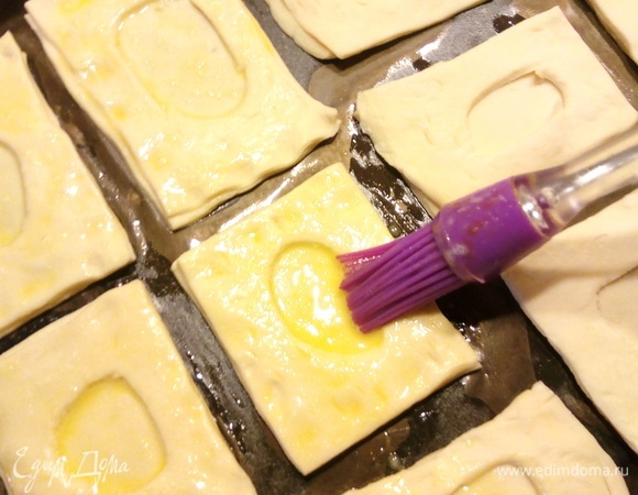 Раскатываем слоеное тесто, нарезаем на прямоугольники произвольной формы, в одном прямоугольнике делаем кружочек посередине и кладем его поверх целого прямоугольника. Так поступаем со всеми. Прямоугольники выкладываем на противень, застеленный пергаментной бумагой, и смазываем взбитым яйцом.