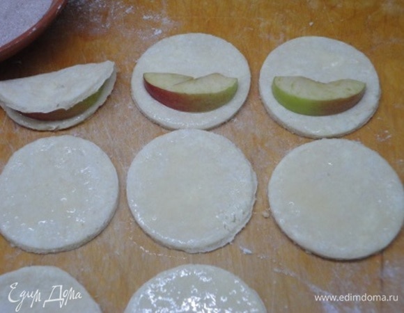 Тесто тонко раскатать на посыпанной мукой поверхности. С помощью стакана вырезать кружочки. Каждый кружочек смазать яйцом и выложить на одну половинку сегмент яблок. Накрыть яблоко второй половиной теста и поверхность теста смазать яйцом.