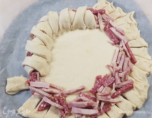 Колбасу нарезать брусочками, сыр натереть на крупной терке. Разложить большую часть колбасы по кругу, заплести полоски теста крест-накрест.