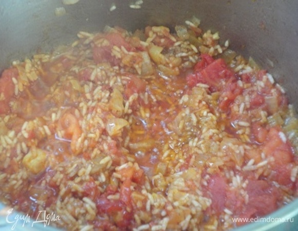 Лук нарезать мелкими кубиками, чеснок порубить. Рис промыть и выложить на кухонное полотенце, чтобы удалить лишнюю влагу. Помидоры измельчить. В кастрюле разогреть масло и обжарить лук до золотистого цвета. Добавить чеснок, перемешать. Сразу же добавить рис, перемешать, затем добавить специи и томаты. Посыпать паприкой и перемешать еще раз. Залить горячим бульоном и варить до готовности риса. Посолить, поперчить.