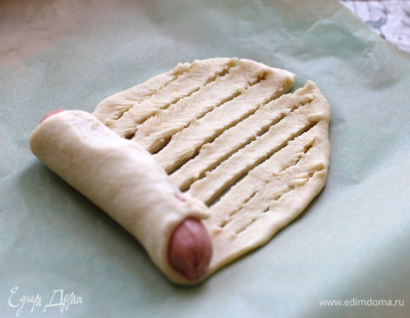 Раскатать шарик в удлиненную лепешку, выложить на один конец очищенную сосиску, далее сделать на тесте продольные надрезы до конца и свернуть сосиску в рулон.