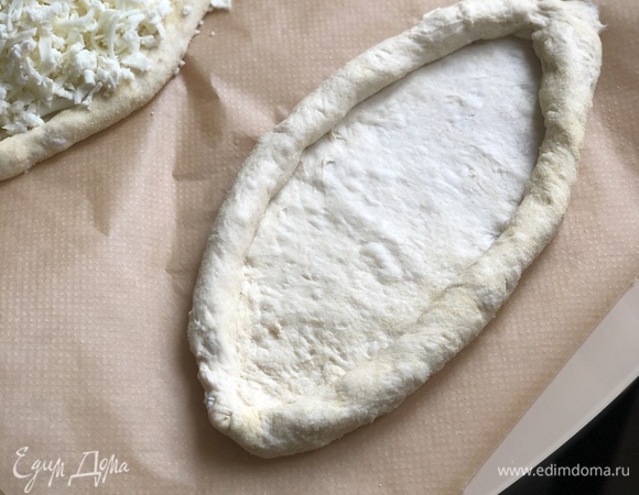 Сформовать хачапури. Для этого рабочую поверхность посыпать семолой (манка из твердых сортов пшеницы), выложить на нее кусочек теста и начать растягивать тесто в достаточно тонкую лепешку. Работать руками, без скалки! Размер лепешки — примерно 20–25 см. Сделать бортики, подкатывая тесто с краев к центру, соединить их в лодочку.