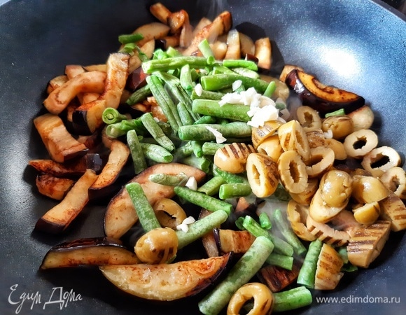 Отправляем стручковую фасоль, рубленый чеснок и оливки, нарзанные колечками, к баклажанам. Солим и добавляем щепотку орегано. Готовим минуты 3 на среднем огне.