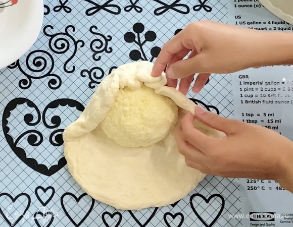 Подошедшее тесто выложить на стол. Руками сформировать небольшой круг-основу для сырного шара. В центр теста выложить сырный шар и собрать тесто вокруг сыра, создавая оболочку сырного шара, защипнуть края теста сверху сырного шара.