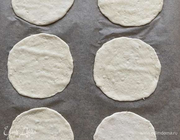 Затем каждый шарик раскатать в тонкую лепешку, переложить на противень, застеленный пергаментом. Накрыть противень пищевой пленкой и дать постоять еще 15–20 минут. В это время разогреть духовку до 260°C. Разогревать не менее 20 минут.