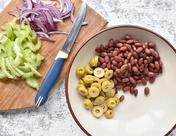 Стебли сельдерея зачистить от грубых волокон и нарезать ломтиками. Красный лук очистить от шелухи и нашинковать полукольцами. В миске соединить отварную красную фасоль и нарезанные колечками оливки. Вместо оливок можно использовать маслины.