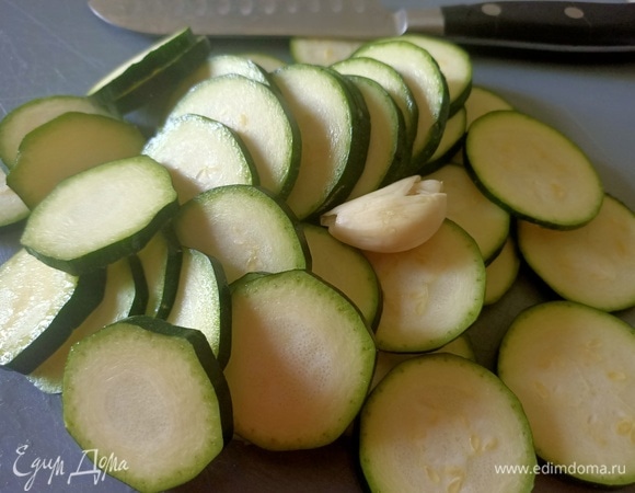 Цукини нарезать кольцами, чеснок раздавить тыльной стороной ножа. В глубокой сковороде разогреть оливковое масло, выложить цукини с чесноком и жарить в течение 2–3 минут.