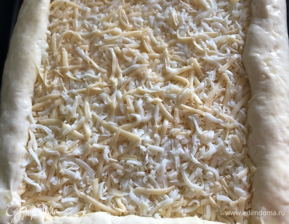 Тем временем натираем сыр на крупной терке. Когда тесто подошло, раскатываем его в прямоугольник, выкладываем на противень, посыпаем тертым сыром, края скручиваем в рулет к центру и защипываем в уголках. Яйца немного взбиваем и хорошо смазываем края нашего пирога, остатки яиц выливаем в центр. Накрываем пирог планкой и оставляем на 20–30 минут подходить.
