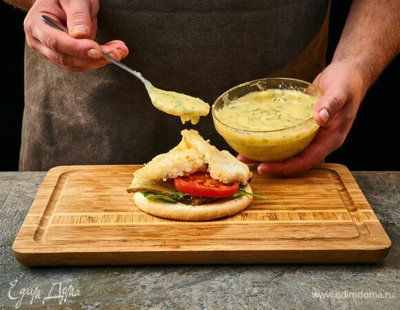 Нижнюю половину булочки полейте соусом, выложите листья шпината, сверху — помидоры, лук и соленые огурцы, далее — обжаренную рыбу и соус.