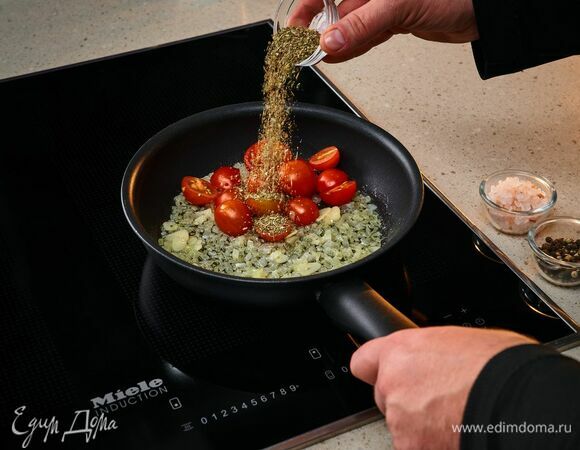 На индукционную панель конфорок Miele поставьте сковороду, добавьте оливковое и сливочное масло, обжарьте лук и чеснок до мягкости. Добавьте черри и орегано. Посолите, поперчите. Протушите 5 минут.