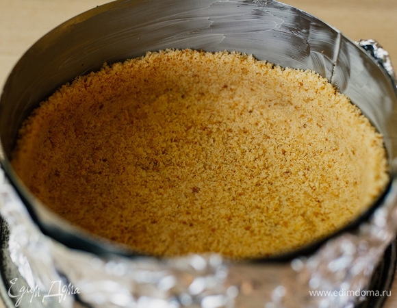 У меня кольцо диаметром 18 см, его дно необходимо выстелить пергаментом, зафиксировать пищевой фольгой и поставить на какую-то форму с большим диаметром, чтобы чизкейк было удобно переносить из духовки. Дно и бортики кольца смазать сливочным маслом. Выложить и разровнять песочную крошку с помощью дна стакана.