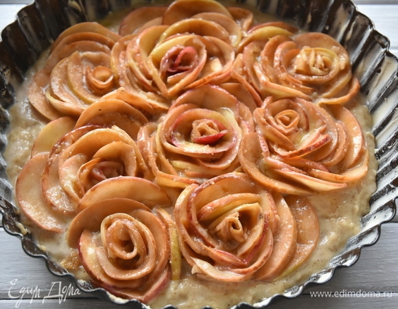 Форму для выпечки смазать сливочным маслом. Посыпать небольшим количеством муки, излишки стряхнуть. Переложить тесто в форму. Из яблочных ломтиков свернуть заготовки цветов и распределить их по поверхности теста. Оставшиеся яблоки распределить вокруг свернутых заготовок, имитируя лепестки роз. Растопить в микроволновой печи оставшееся сливочное масло и густо смазать яблочные цветы. Сверху посыпать тростниковым сахаром. Выпекать яблочный пирог в духовке, разогретой до 180°C, около одного часа.