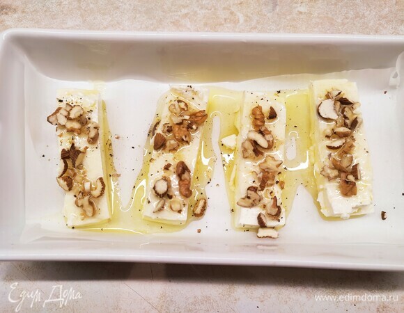 Фету выложить в форму для запекания. Можно целым куском, а можно порционно нарезать. Смешать оливковое масло и мед до однородности. Вылить смесь на фету, посыпать орехами и поставить в духовку на 15 минут при температуре 200°C.