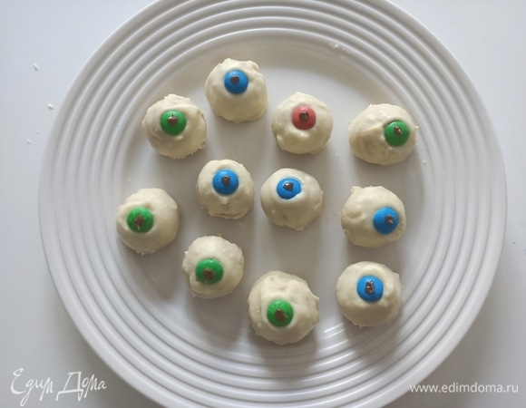 Достать конфеты из холодильника и покрыть их белым шоколадом. В центр уложить цветную конфету и капнуть маленькую каплю темного шоколада. Убрать в холодильник.