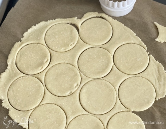 Муку смешать с солью и разрыхлителем, просеять. Холодное масло натереть на терке, добавить холодный творог и быстро замесить тесто. Чем меньше будет контакта с теплыми руками, тем лучше. Я все ингредиенты складываю в комбайн и замешиваю тесто без контакта с телом в течение 1 минуты. Разделить тесто на 2 части: с одной работаем, другую кладем в холодильник. Чтобы не забивать тесто мукой, можно взять 2 листа пергамента и раскатать между ними тонкий пласт, вырезать круги (у меня диаметр — 6 см).