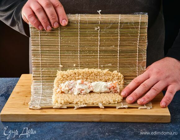 Ровным слоем выложите рис на бамбуковый коврик, обернутый в пленку. В центр рисовой массы положите начинку из рыбы и сыра. Поднимите края коврика и скатайте большой ролл.