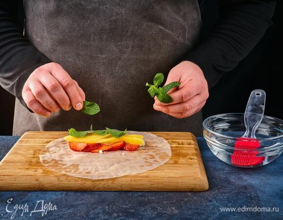 Пластичный лист рисовой бумаги переложите на доску. На него выложите 2 ст. л. творожной начинки, горсть клубнично-манговой нарезки и несколько листиков мяты.